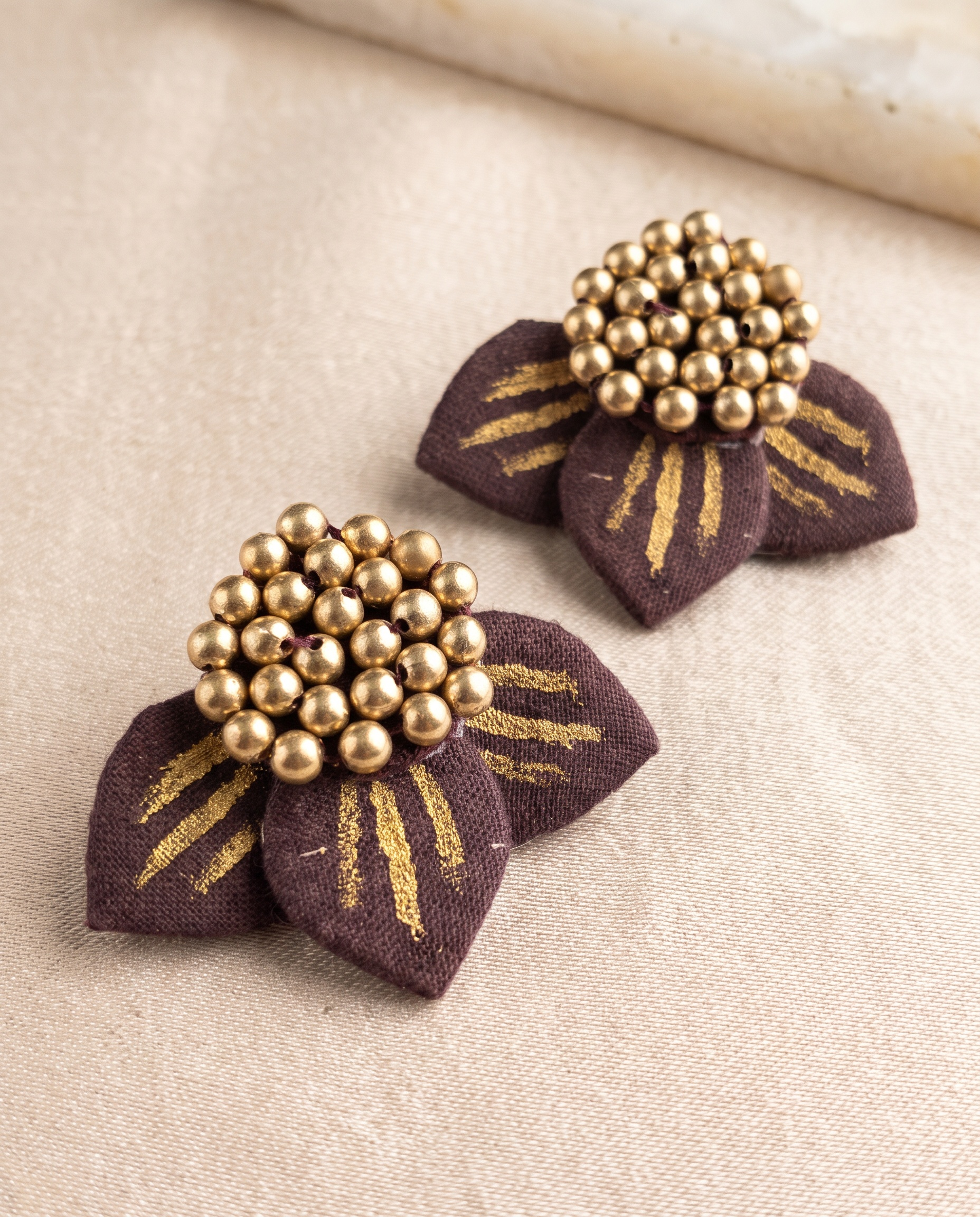 Close-up of maroon fabric floral earrings with gold bead detailing on neutral fabric backdrop