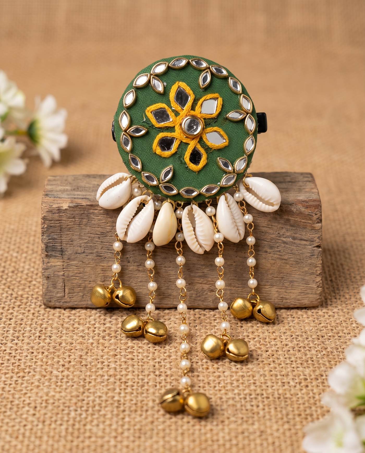 Green mirror hair brooch with shell and bell tassels displayed on wood block with flowers