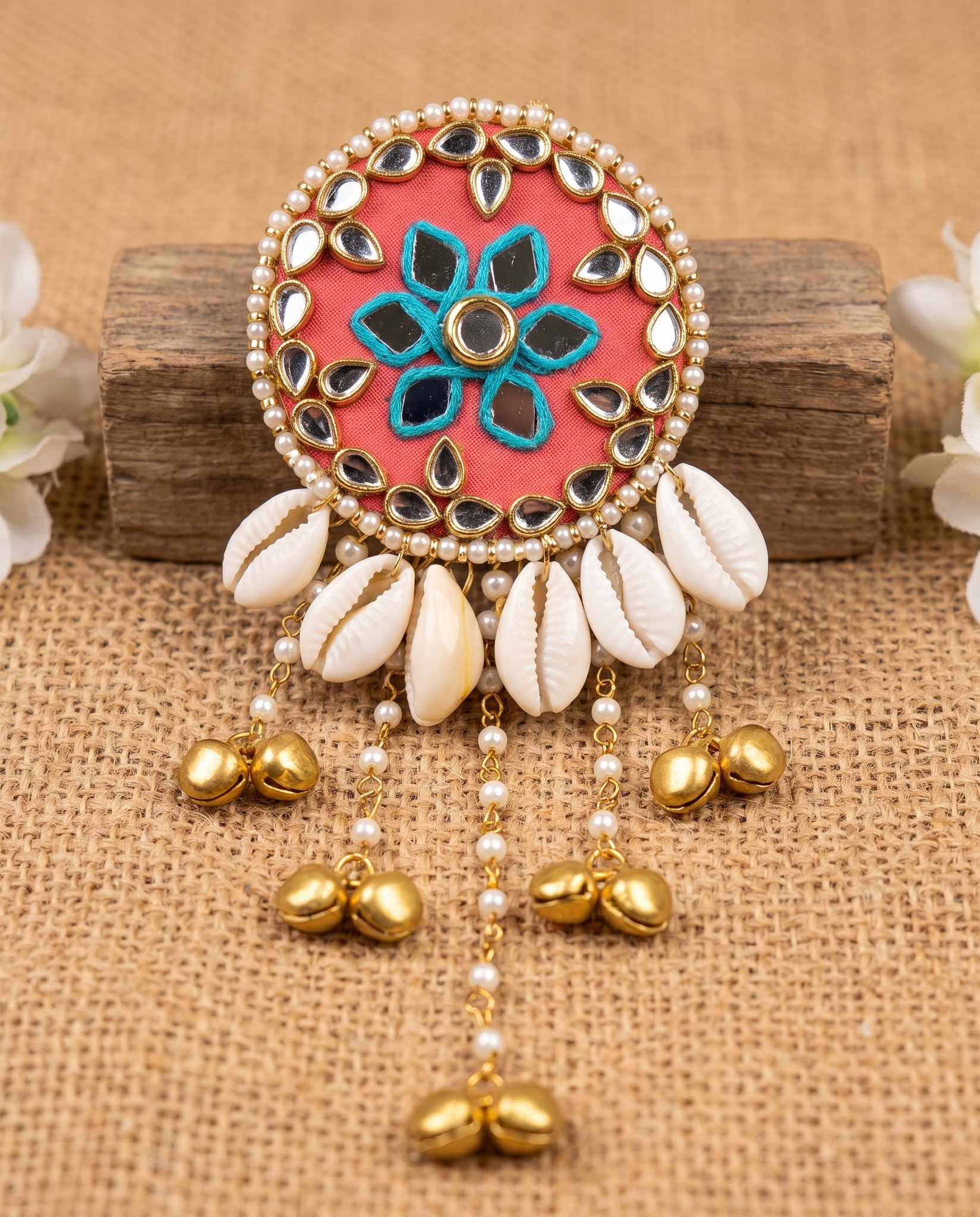 Coral mirror hair brooch with cowrie shells and gold bells displayed on wood block with flowers