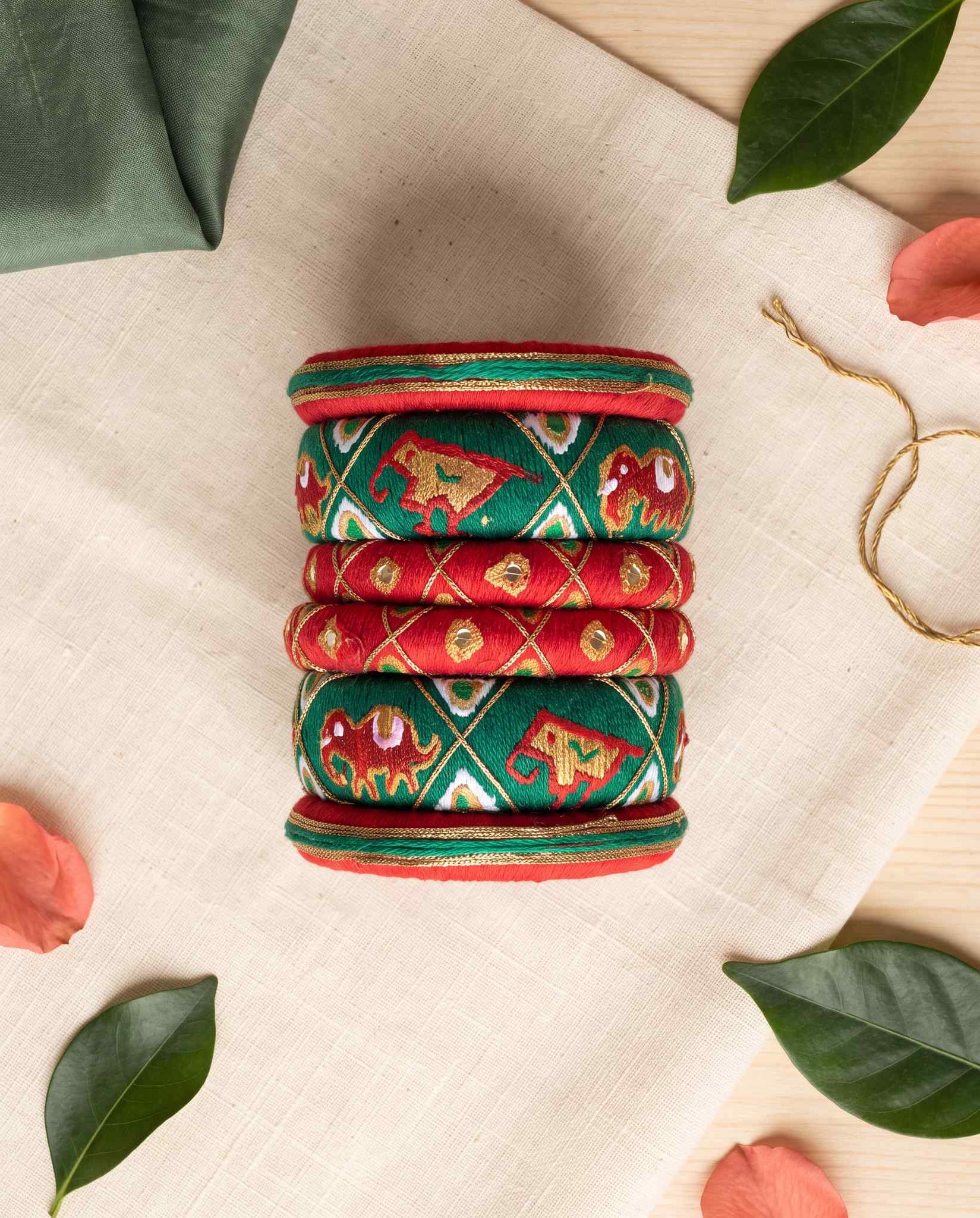 Red and green elephant silk thread bangles styled with leaves and petals on cream linen