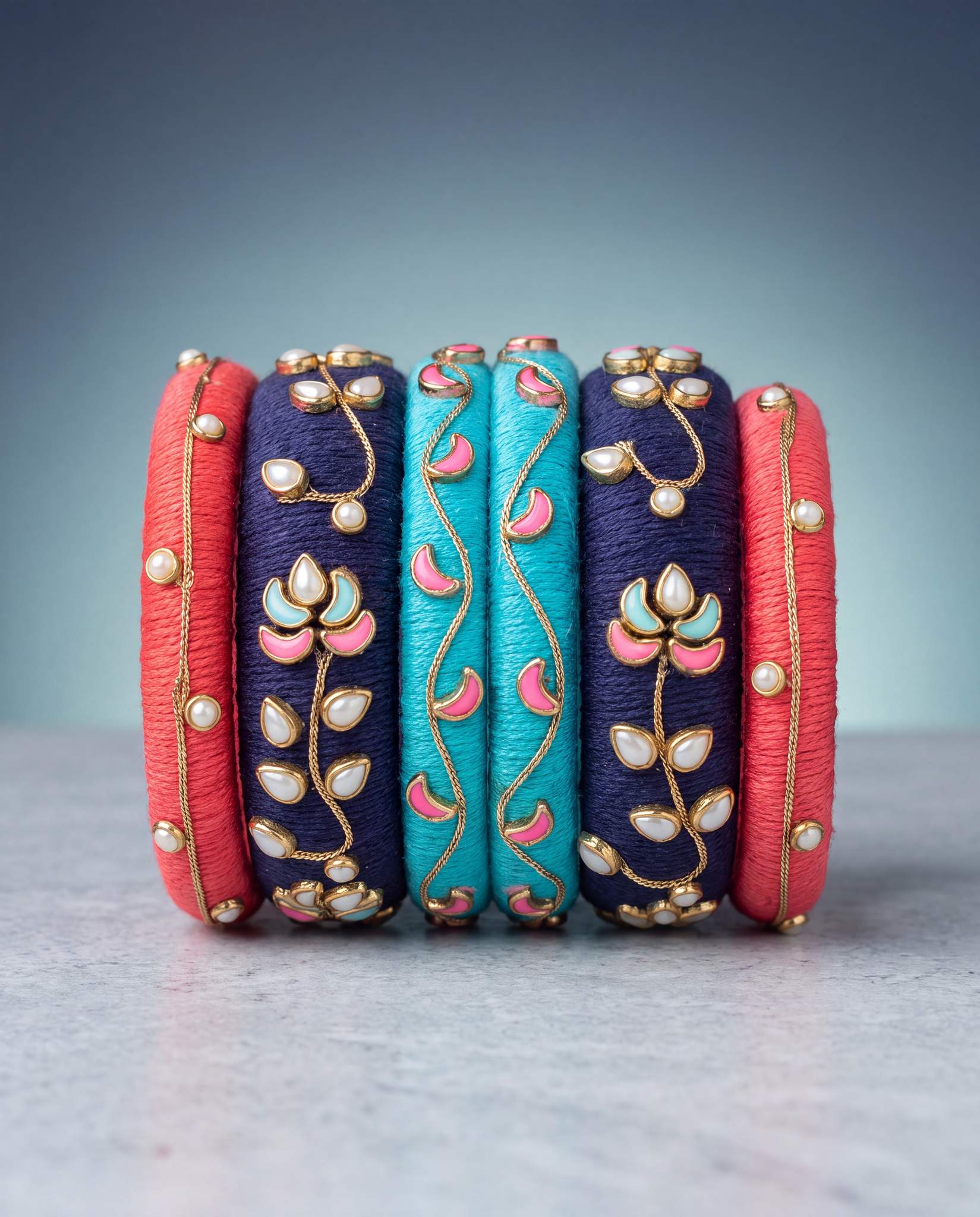 Red, navy, and turquoise silk thread bangles with pearl floral accents on gradient background