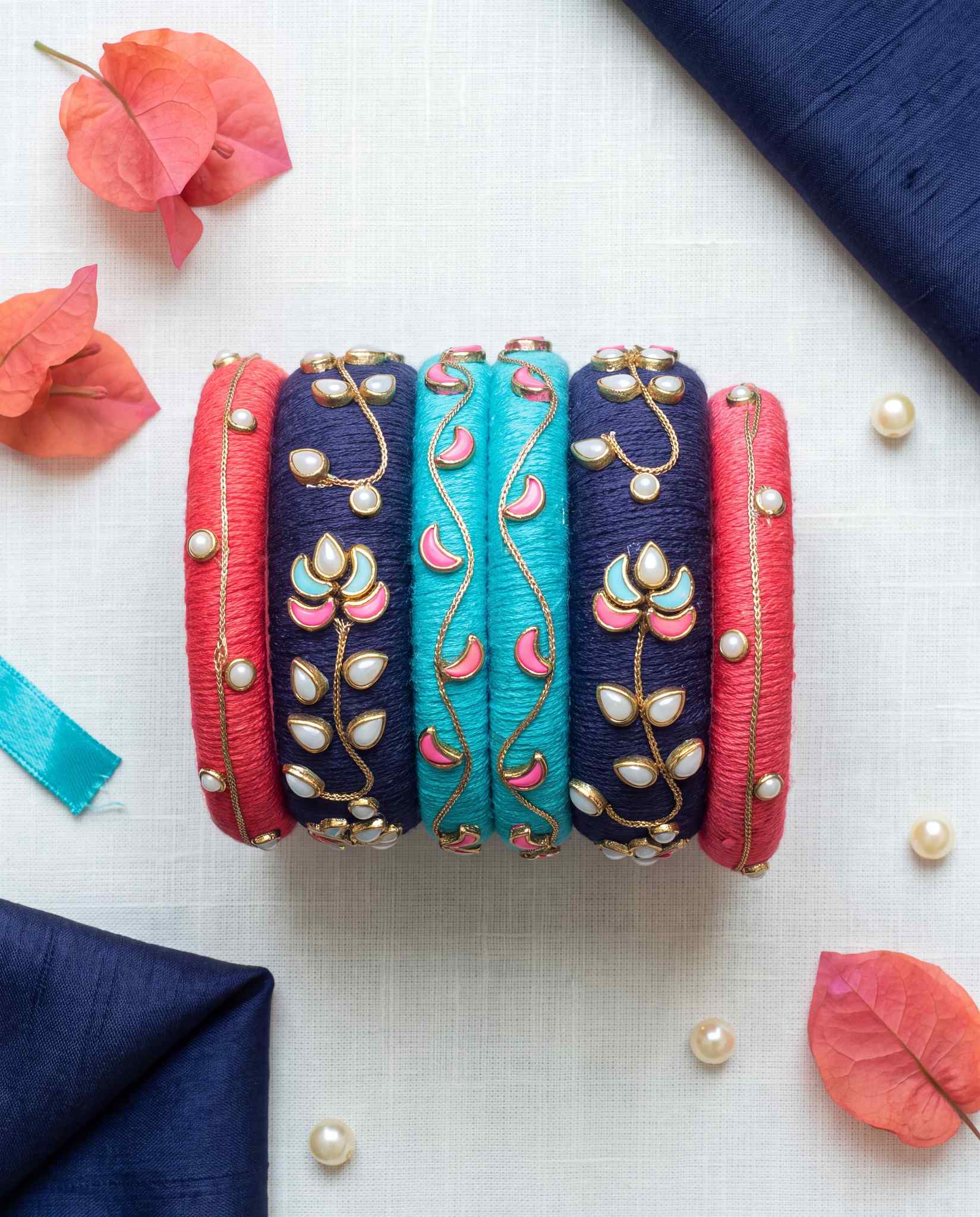 Red, navy, and turquoise meenakari bangles styled with pearls and coral flowers on white linen