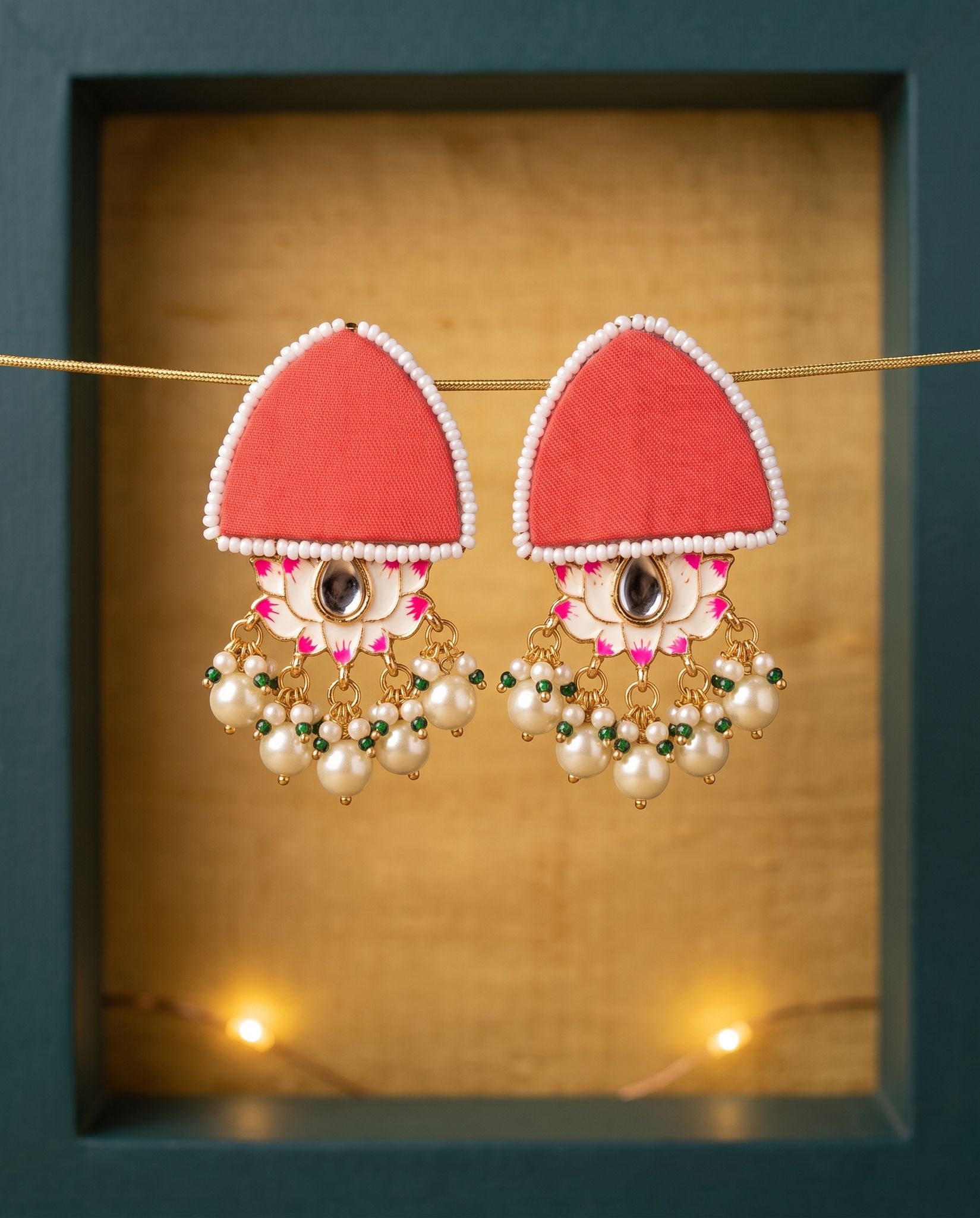 Red pearl floral earrings displayed in teal frame with gold backdrop and candlelight
