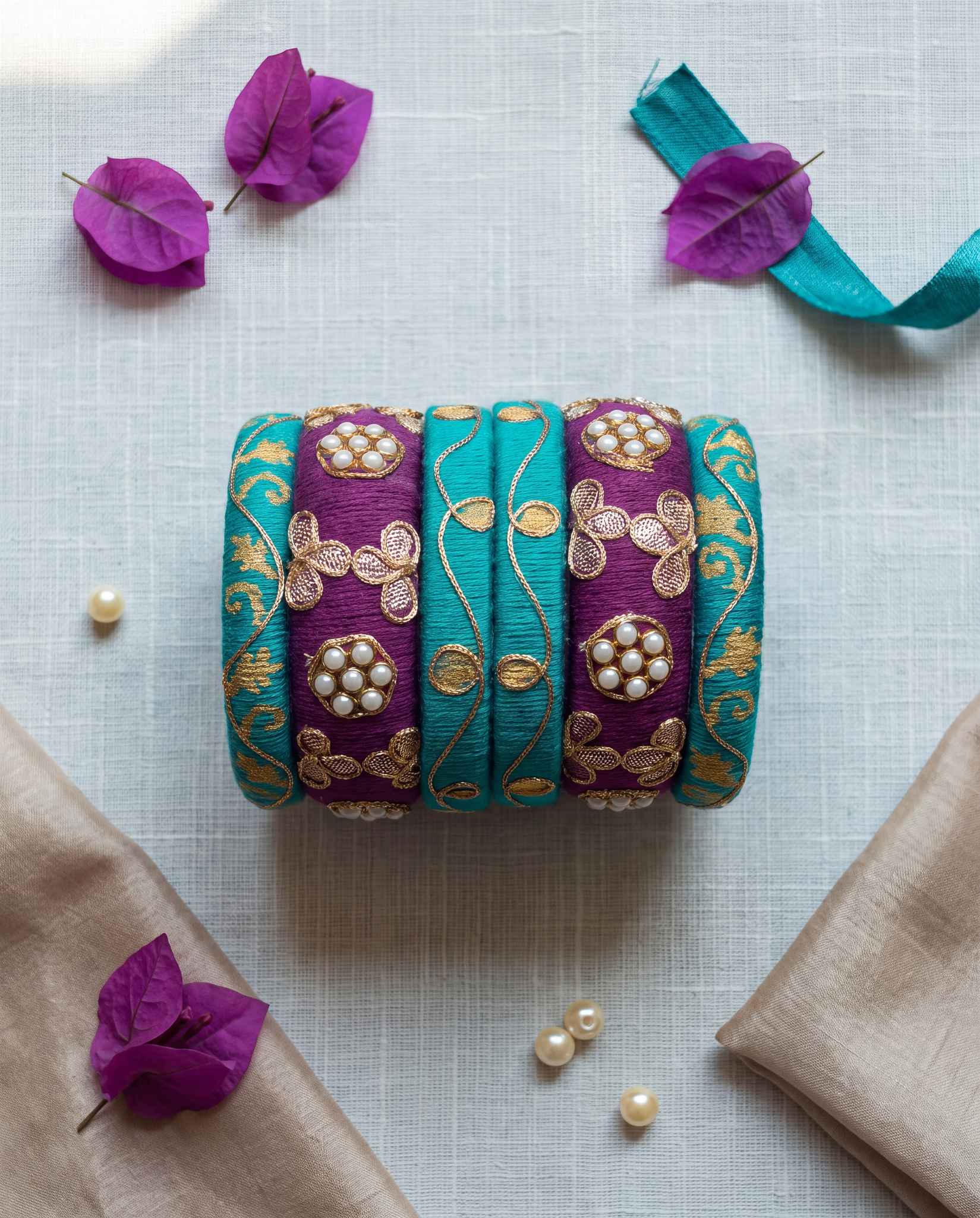 Teal and plum zari kundan bangles styled with purple flowers and pearls on white linen
