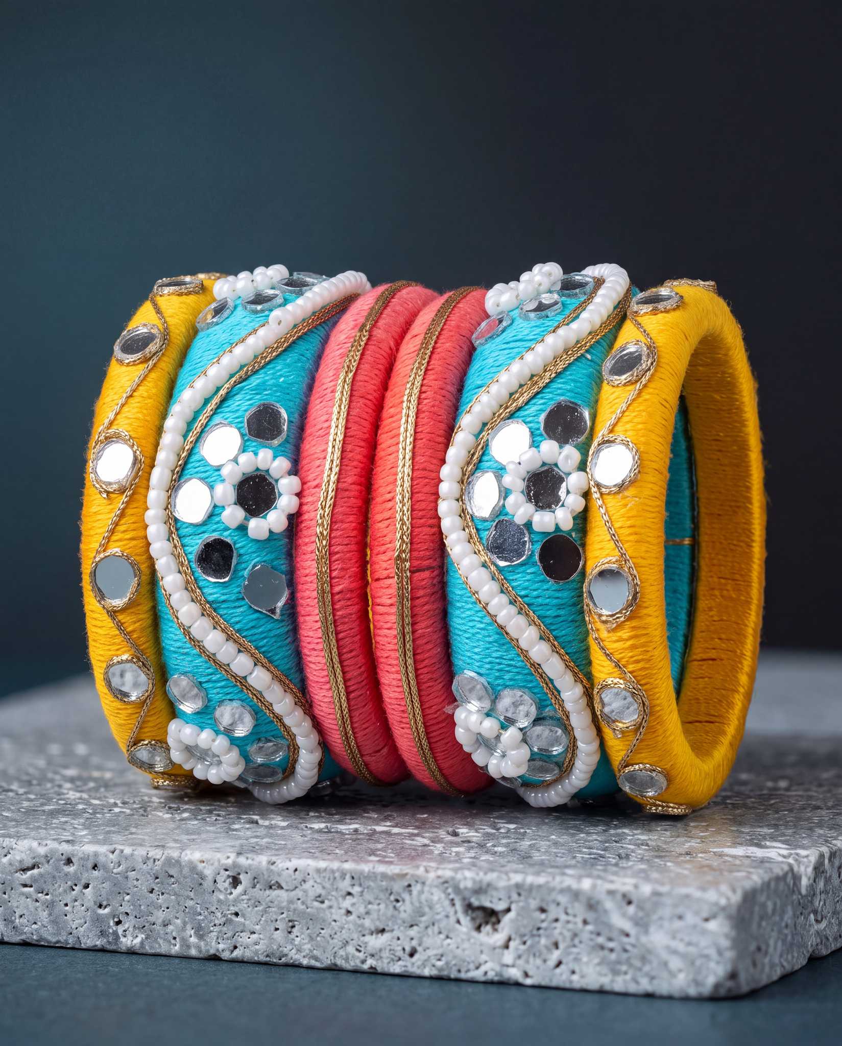 Turquoise, yellow, and pink silk thread bangles with mirror work displayed on dark stone background