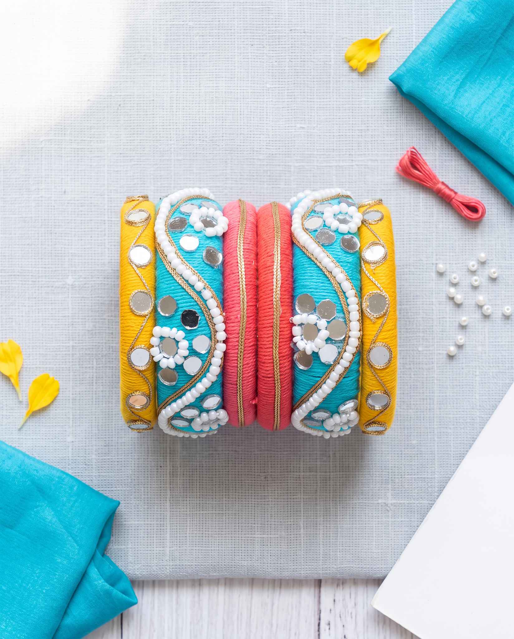 Turquoise, yellow, and pink mirror beadwork bangles styled with yellow petals and pearls on linen