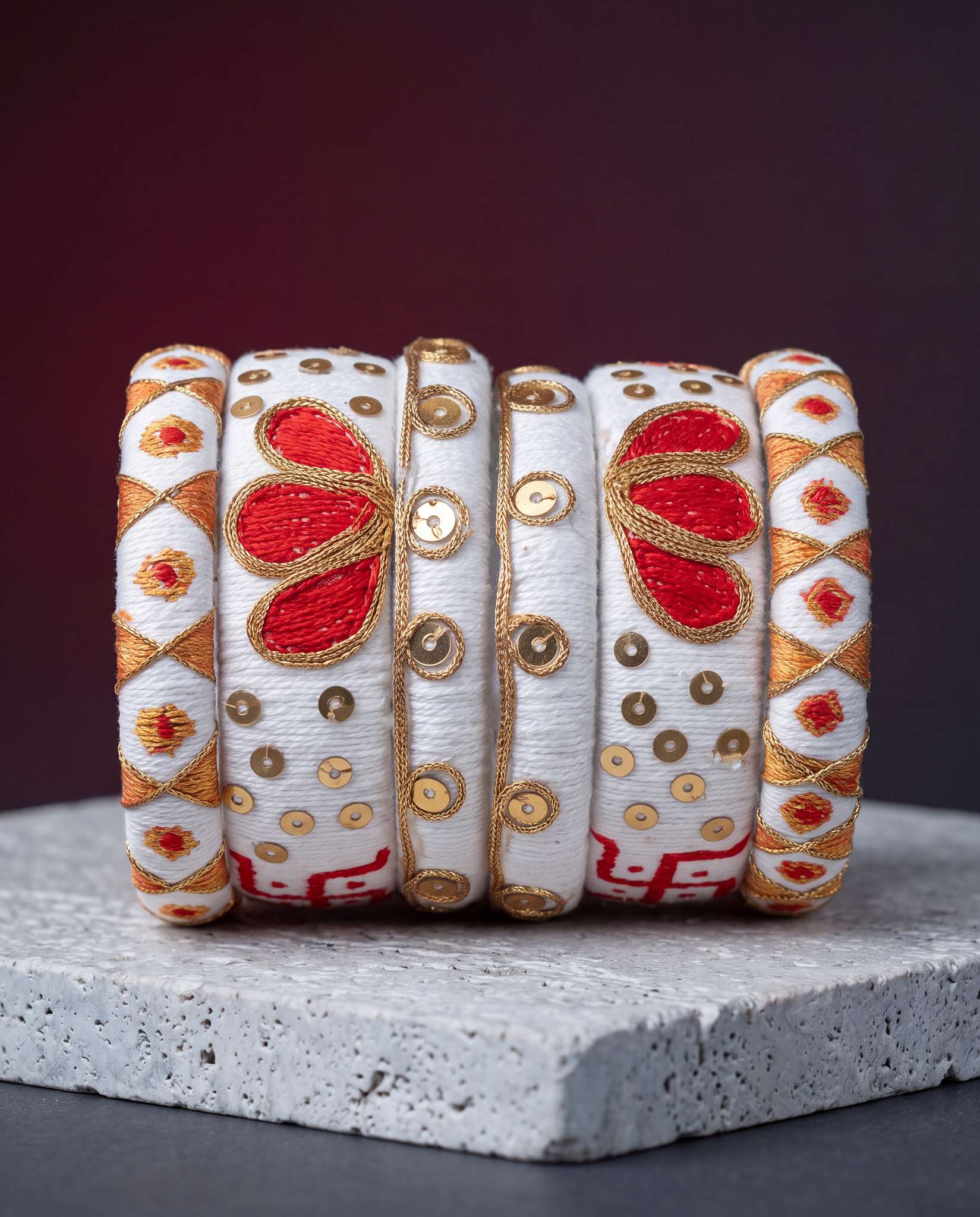 White silk thread bangles with red and gold embroidery displayed against a dark maroon background