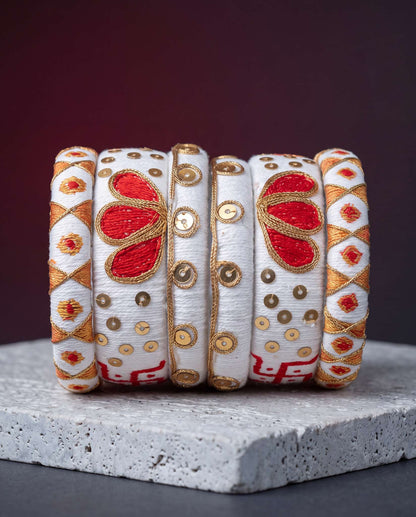 White silk thread bangles with red and gold embroidery displayed against a dark maroon background